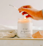 A hand holding a lit match over a glowing candle in a jar labeled "Large Soy Candle - Kiwi & Guava by MyHomeDecor.ca." The white jar sits on a tray beside some stones and a pink crystal, creating a serene and aesthetic composition. With tropical and fruity scents like agave, ananas, and lime, this environmentally friendly candle is perfect for relaxation.