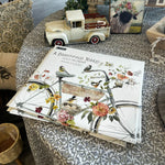 A 2025 Calendar A Beautiful Ride from MyHomeDecor.ca rests on a table, featuring a floral bicycle illustration on its linen embossed paper cover. Surrounding it are decorative items including a vintage toy truck, a framed picture of an animal, a wicker basket, and cushions with intricate patterns.