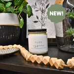 A Large Soy Candle - Yuzu and Cypress by MyHomeDecor.ca is displayed on a black surface with a wooden bead garland, small dishes, and a plant in the background. The soy candle is in a clear jar with a black lid and offers 60 hours burn time. A green text bubble on the right side says "NEW!".