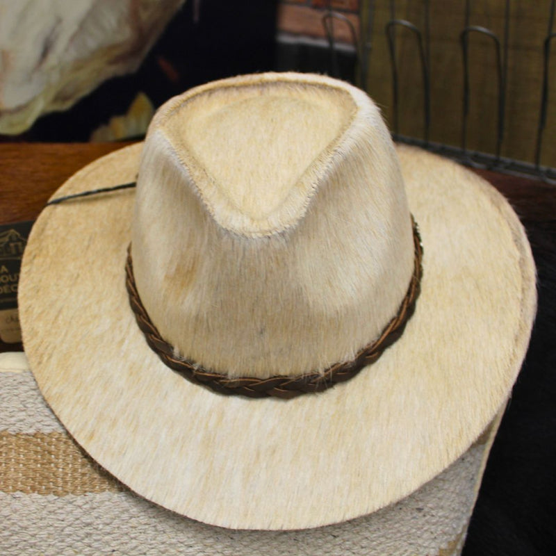 A MyHomeDecor.ca Cowboy Hat Blond Small with a braided brown band around the base and a crease down the crown sits on a textured surface. The background is out of focus, displaying some other objects and a wire-frame structure, showcasing an authentic touch of Western fashion.