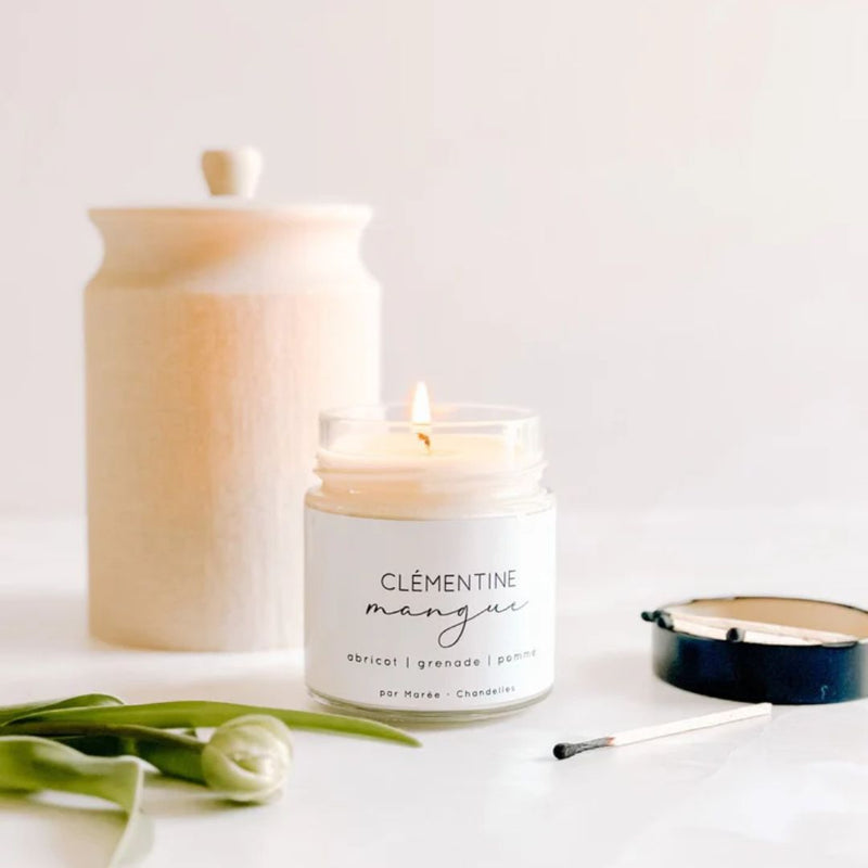 A lit Medium Soy Candle Clementine & Mango from MyHomeDecor.ca in a glass jar sits on a white surface next to a wooden container with a lid, an open matchbox with a used match, and a green tulip stem.