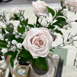 A floral arrangement of pale pink roses is surrounded by small white flowers and green leaves, displayed in an indoor setting on a table. One 27" Fresh Touch Vienna Rose Spray - Blush by MyHomeDecor.ca in the center is prominently in focus, showing detailed petals and soft coloration.