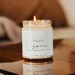 A lit Large Soy Candle - Yuzu and cypress in a glass jar sits on a reflective surface. The label reads, "Yuzu Cyprès, poire | ylang ylang | néroli, par MyHomeDecor.ca." Crafted from soy wax and offering a 60 hours burn time, the softly blurred background suggests a cozy indoor setting.