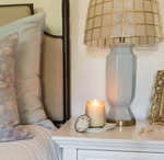 A cozy bedroom scene with a light blue and beige pillow on a bed. A white nightstand holds a lit Large Signature Candle - Magnolia by MyHomeDecor.ca in a decorative holder, a small clock, a blue lamp with a gold grid-patterned lampshade, and a small ornamental gold frame.