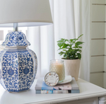 A bedside table features a blue and white patterned lamp, a small potted plant, a lit Large Signature Candle - Magnolia from MyHomeDecor.ca, and a textured hobnail jar sitting on a stack of books, all set against a backdrop of light-colored curtains and walls.