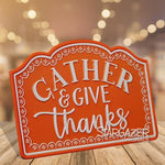 An orange and white metal table sign with scalloped edges displays the message "Gather & Give Thanks" in white text. Placed on a wooden surface, it is complemented by a blurred background of warm, ambient lights. The word "MyHomeDecor.ca" is visible at the bottom right, perfect for fall table decor.