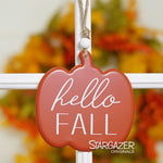 A red pumpkin-shaped metal sign hangs on a white cross-shaped structure, proudly displaying the phrase "hello FALL" in elegant white cursive. In the background, autumn leaves are blurred, adding to the seasonal charm. The name "MyHomeDecor.ca" is printed at the bottom right of the image.