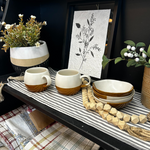 A display shelf features a potted plant with white and green flowers, the Ball Mug - 2 Tone from MyHomeDecor.ca, stacked bowls, and a botanical print in the background. The items rest on a striped cloth with a wooden bead garland draped across the shelf, adding a rustic touch.