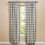 A pair of rustically charming curtains in a bold plaid print of gray, white, and faint yellow hang on a black curtain rod, framing a window with greenery outside. The beige walls and light wood floor enhance the farmhouse aesthetic. The **Curtain - Limestone Lined Panel Pair** by **MyHomeDecor.ca** perfectly complements the decor.
