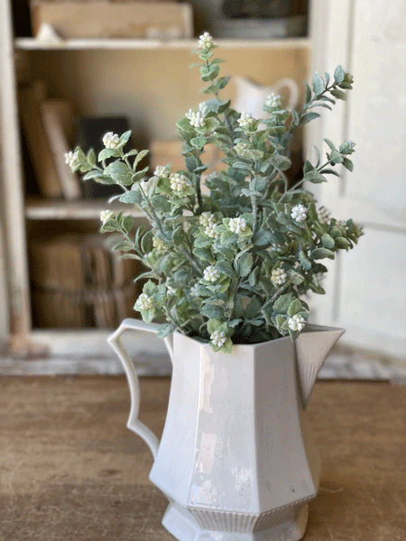 A white geometric pitcher, **Bush - Meadow Whisper** by **MyHomeDecor.ca**, is used as a vase, holding lush sage green flowers and small white blossoms. The pitcher sits on a wooden surface, with a blurred background showcasing shelves filled with stacked books and decor items.