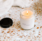 A lit candle in a small glass jar labeled "MyHomeDecor.ca Large Soy Candle - Lavender" sits on a terrazzo countertop with scattered earthy tones, its black lid placed nearby. Infused with lavender essential oils, this indoor & outdoor candle creates a serene ambiance. A soft, white cloth is in the background, slightly out of focus.