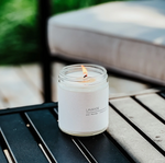 A lit Large Soy Candle - Lavender by MyHomeDecor.ca in a glass jar labeled "Lavande," infused with lavender essential oils, sits on a black metal table. The background features an out-of-focus light beige cushion and greenery, suggesting an outdoor setting. The scene evokes a calm and relaxing atmosphere.